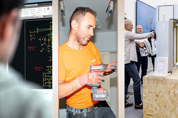 Person in orangefarbener Arbeitskleidung montiert mit einem Akkuschrauber ein Fensterelement in einer Schulungs- oder Werkstattumgebung, während im Hintergrund technische Pläne und Demonstrationsmodelle zu sehen sind.